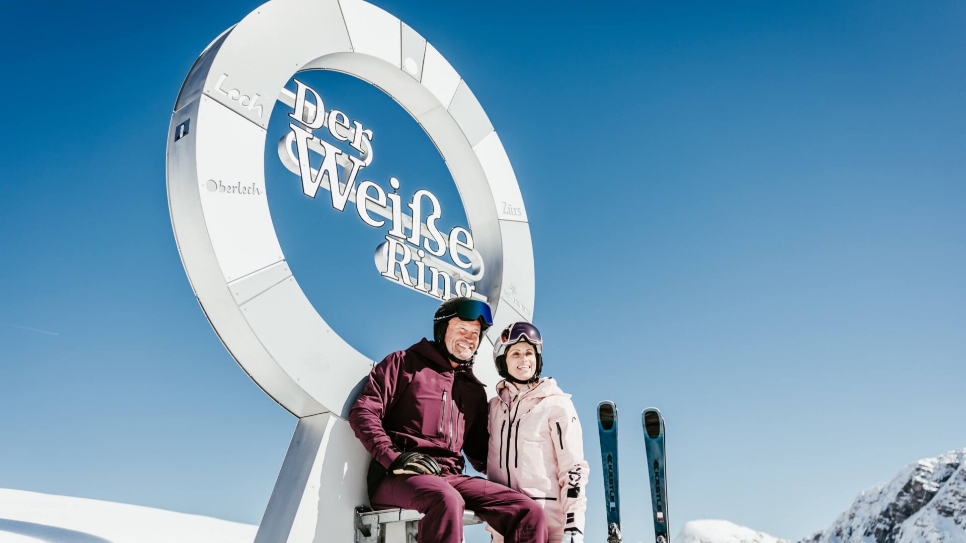 Skifahrer-Paar sitzt am weißen Ring in Lech/Zürs mit herrlichem Bergpanorama und blauen Himmel im Hintergrund.