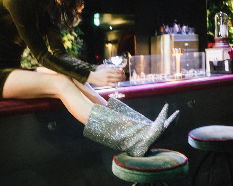 Frau im glitzernden Outfit sitzt an der Bar in einem stilvollen Hotel, genießt ihren Drink in trendiger Atmosphäre.