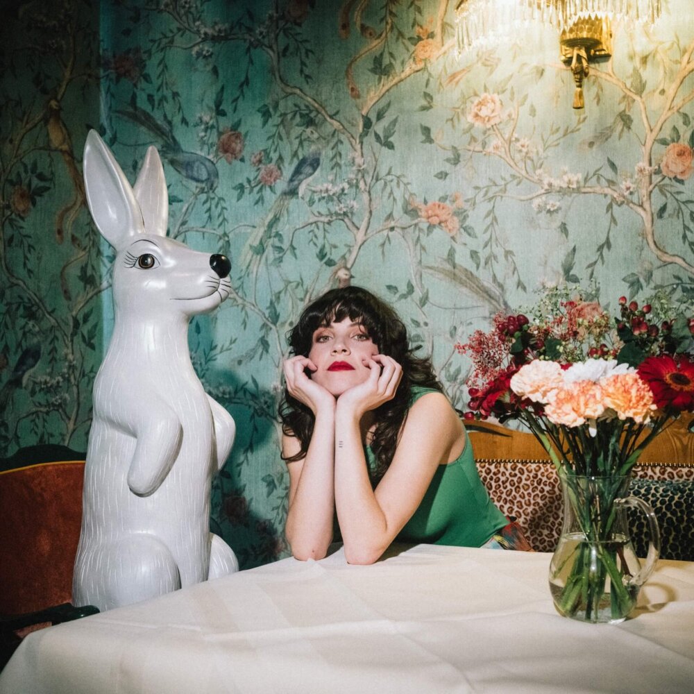 Frau mit roten Lippen sitzt am Tisch neben einem weißen Hasen und bunten Blumen in einem stilvollen Restaurant.