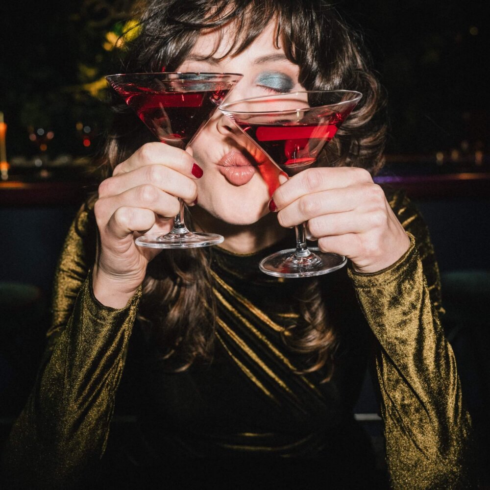Frau mit glamourösem Outfit hält zwei Cocktails in der Bar des Hotels Edelweiss in Lech/Zürs. Sie lächelt fröhlich.