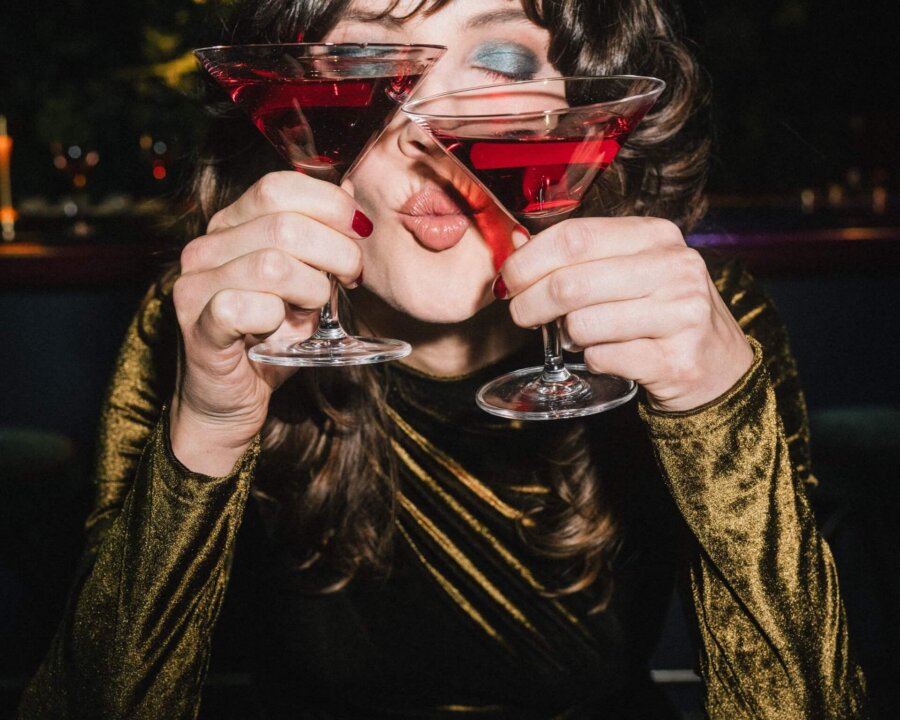 Frau mit glamourösem Outfit hält zwei Cocktails in der Bar des Hotels Edelweiss in Lech/Zürs. Sie lächelt fröhlich.