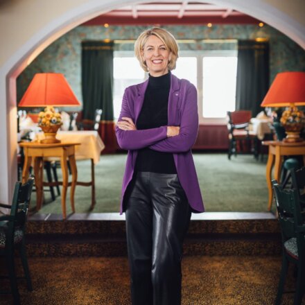 Lächelnde Frau in lila Blazer steht im stilvollen Restaurant des Hotels Edelweiss in Lech/Zürs. Gemütliche Atmosphäre.