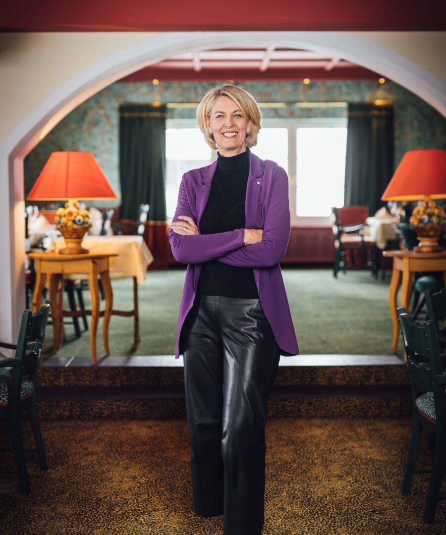 Lächelnde Frau in lila Blazer steht im stilvollen Restaurant des Hotels Edelweiss in Lech/Zürs. Gemütliche Atmosphäre.