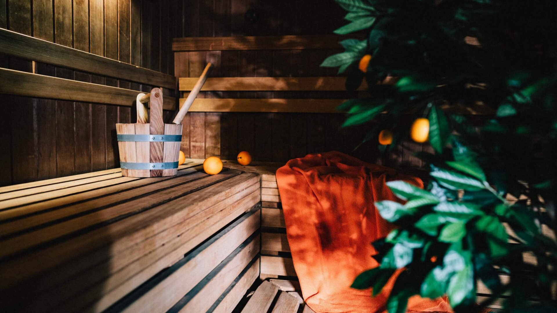 Gemütliche Sauna im Hotel Edelweiss mit Holzbank, Aufgusszubehör und einem orangefarbenen Handtuch zur Entspannung.