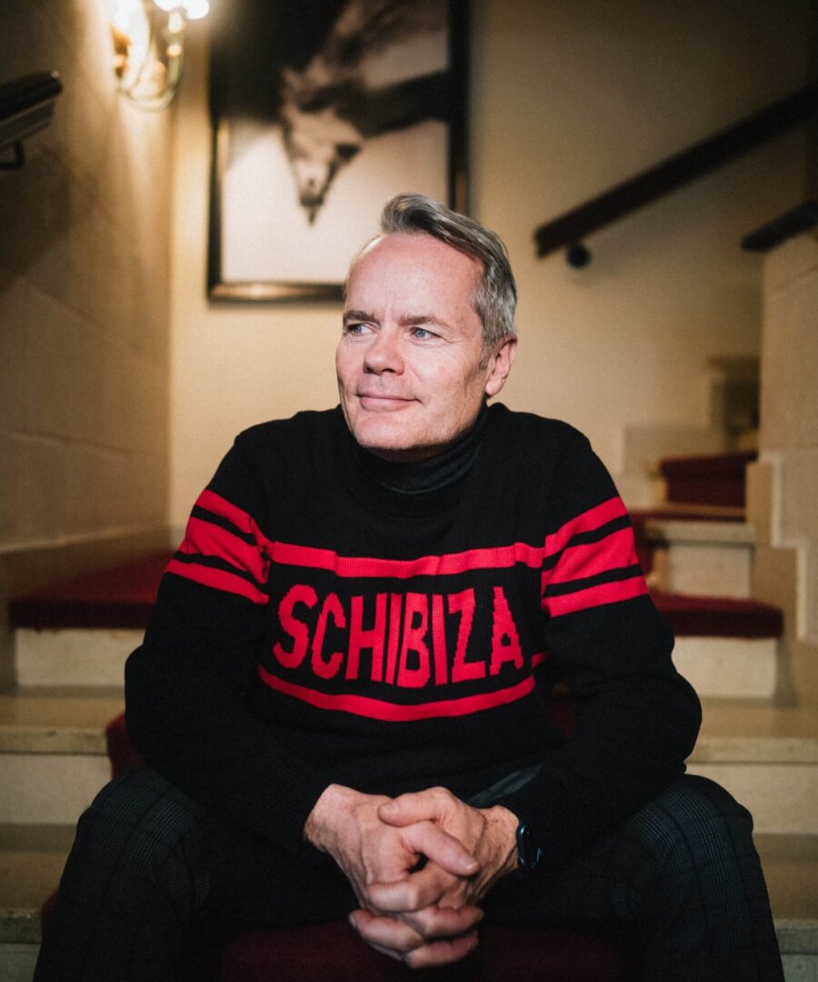 Mann sitzt auf einer Treppe im Hotel Edelweiss, trägt einen schwarzen Pullover mit rot gestreiftem Schriftzug.