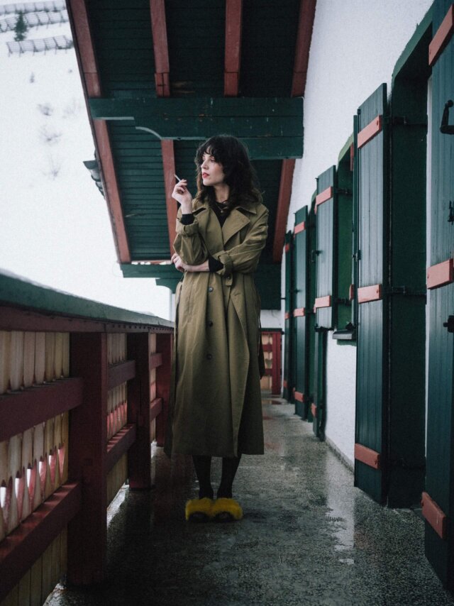 Frau in einem beigen Trenchcoat und gelben Hausschuhen auf dem Balkon eines Hotels in den verschneiten Alpen.
