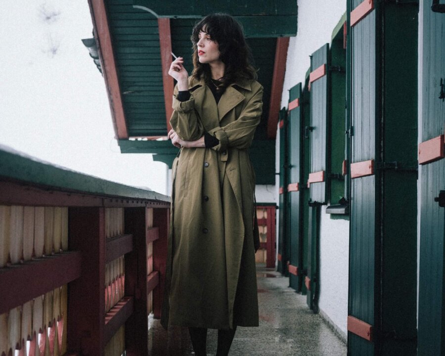 Frau in einem beigen Trenchcoat und gelben Hausschuhen auf dem Balkon eines Hotels in den verschneiten Alpen.
