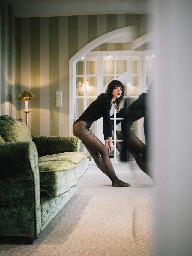 Elegante Frau in einem stilvollen Hotelzimmer im Hotel Edelweiss, Lech/Zürs, mit einem grünen Sofa und Spiegel.