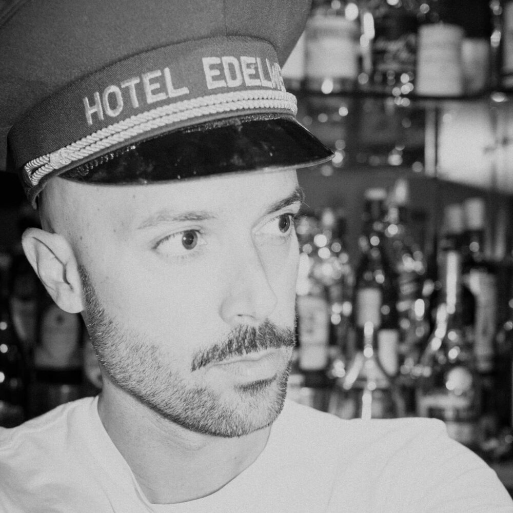 Mitarbeiter des Hotel Edelweiss trägt seine charakteristische Mütze, vor einer Bar mit verschiedenen Spirituosen.