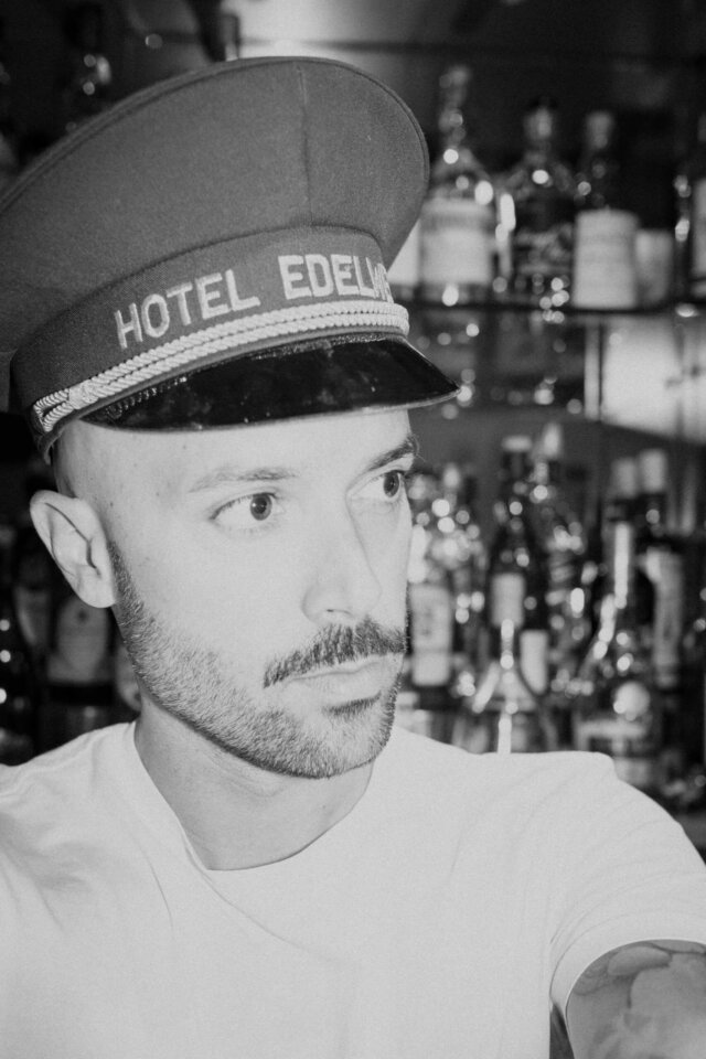 Mitarbeiter des Hotel Edelweiss trägt seine charakteristische Mütze, vor einer Bar mit verschiedenen Spirituosen.