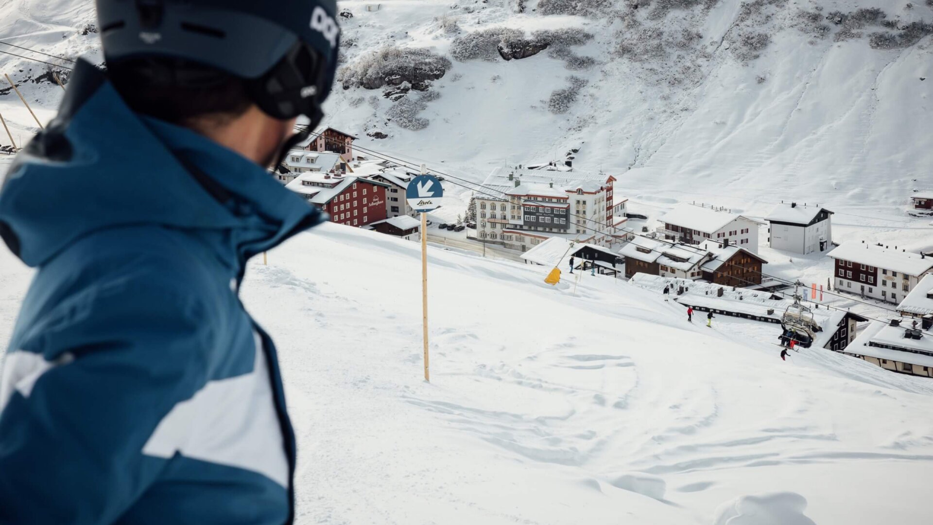 Skifahrer blickt auf die verschneite Landschaft von Lech/Zürs mit dem Hotel Edelweiss und der Seilbahn im Hintergrund.