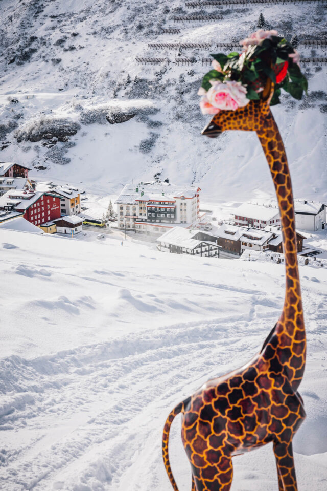 Bunte Giraffe mit Blumenkranz vor verschneiter Alpenlandschaft und Hotel Edelweiss in Lech/Zürs am Arlberg.