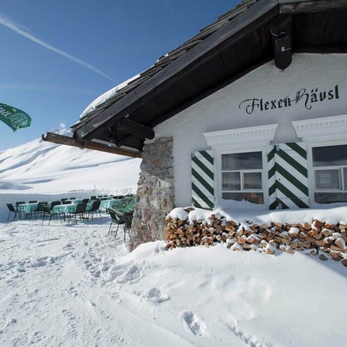 Das Flexen Häusl im Schnee mit sonniger Terrasse und umzäuntem Holzstapel in Lech/Zürs am Arlberg.