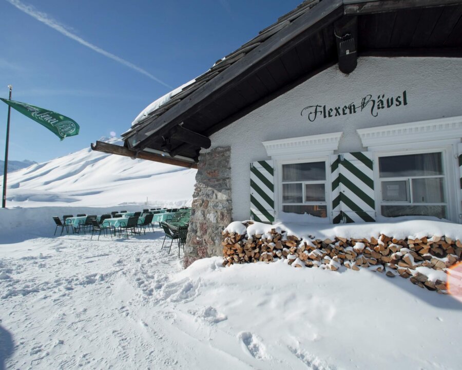 Das Flexen Häusl im Schnee mit sonniger Terrasse und umzäuntem Holzstapel in Lech/Zürs am Arlberg.
