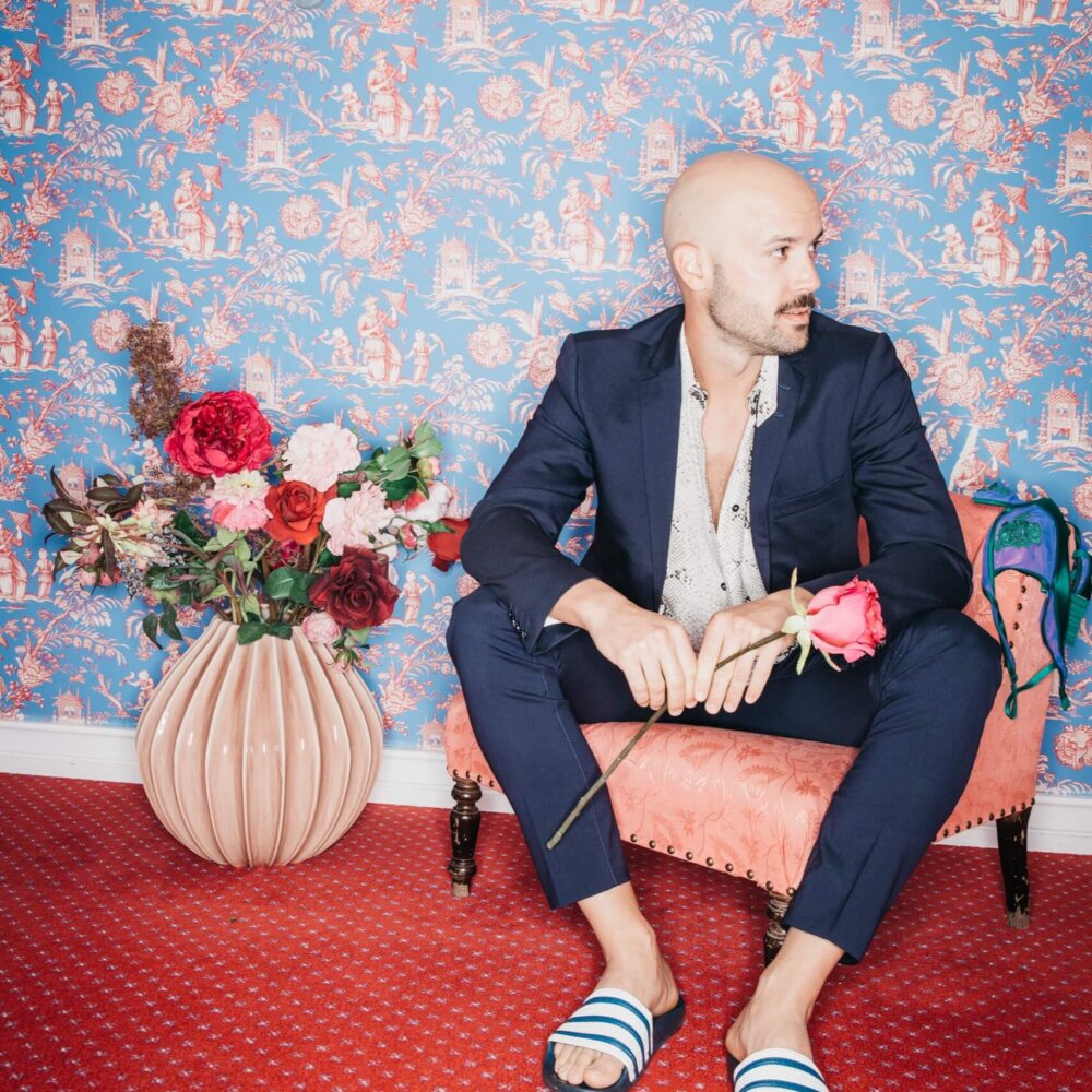 Mann in stilvollem Outfit und Slipper sitzt auf einer pinken Couch vor einer blauen Tapete mit floralen Mustern.