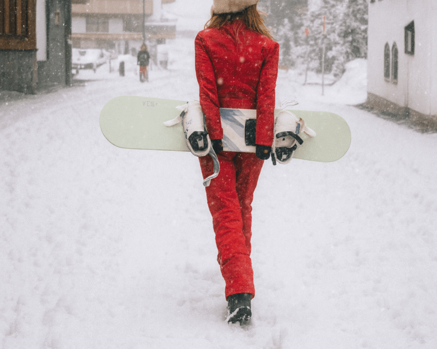 Frau im roten Schneeanzug mit Snowboard spaziert durch die verschneiten Straßen von Lech/Zürs am Arlberg.