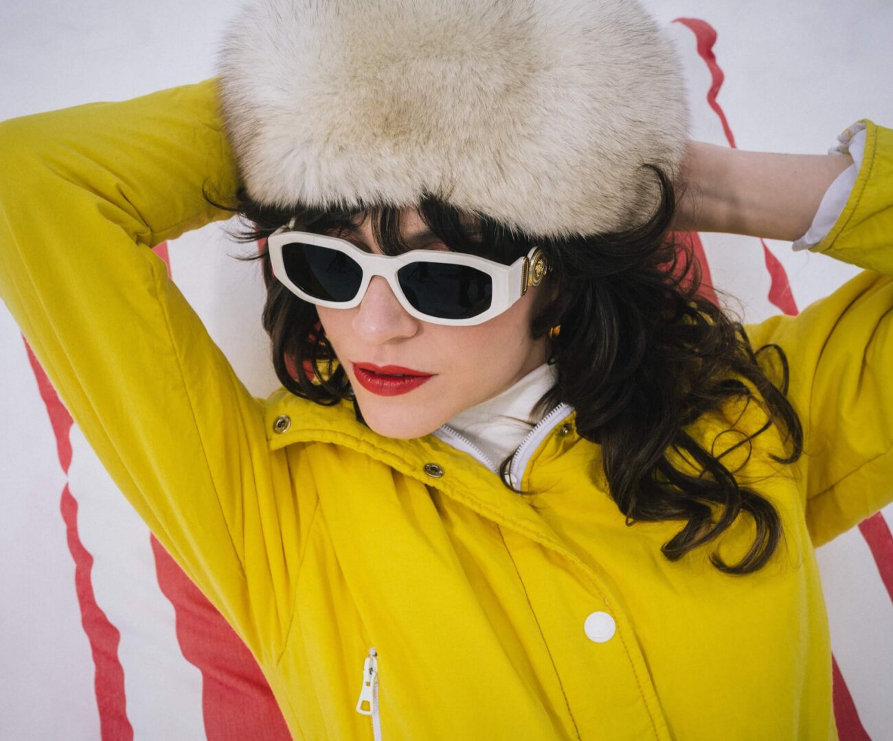 Modische Frau in gelber Jacke und Sonnenbrille, entspannt in Lech/Zürs, umgeben von schneebedeckter Landschaft.