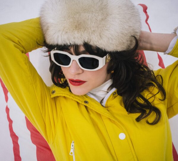 Modische Frau in gelber Jacke und Sonnenbrille, entspannt in Lech/Zürs, umgeben von schneebedeckter Landschaft.