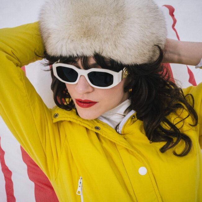 Modische Frau in gelber Jacke und Sonnenbrille, entspannt in Lech/Zürs, umgeben von schneebedeckter Landschaft.