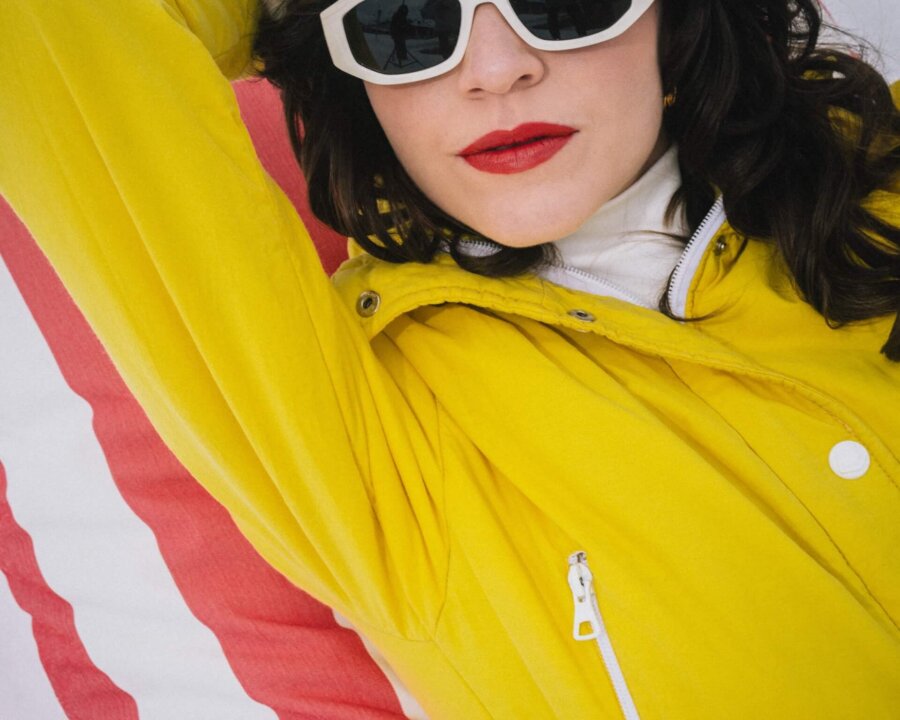 Frau in gelber Winterjacke und Sonnenbrille entspannt auf einer Liege im Schnee, Hotel Edelweiss in Lech/Zürs im Hintergrund.