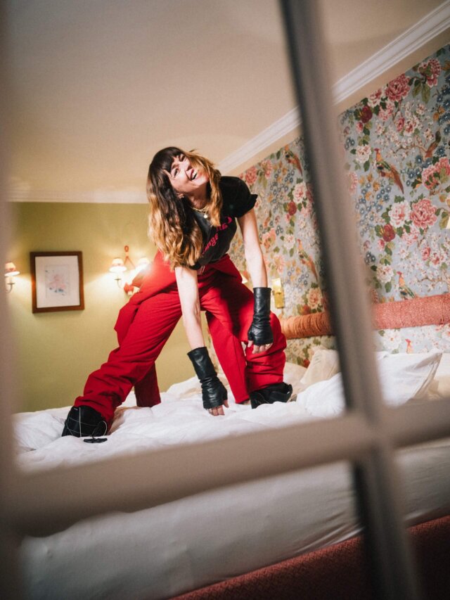 Frau in rotem Outfit posiert auf einem Bett im Hotel Edelweiss, umgeben von stilvollem, floralem Interieur.