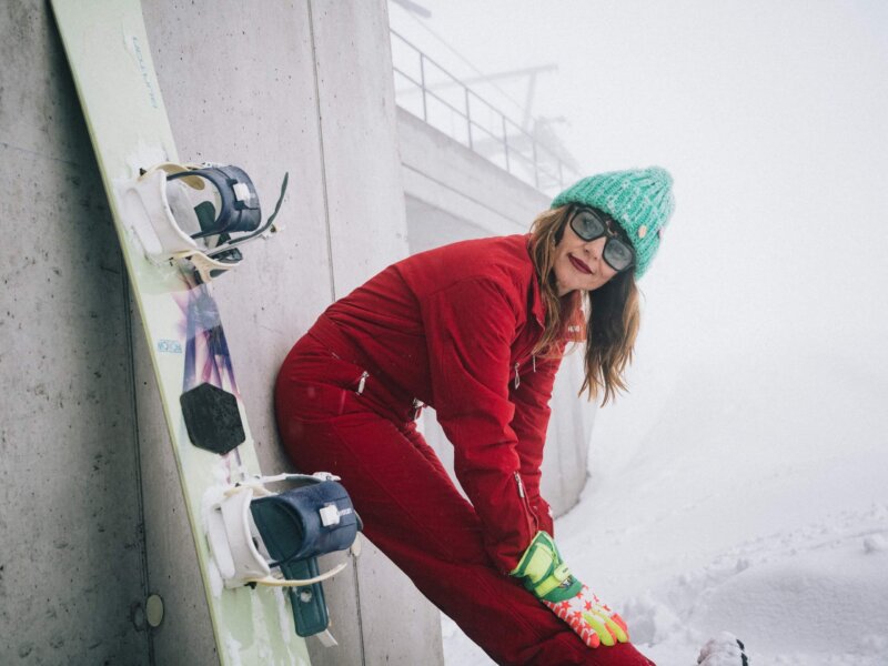 Frau in rotem Skianzug und grüner Mütze bereitet sich zum Skifahren in Lech/Zürs am Arlberg vor, umgeben von Schnee.