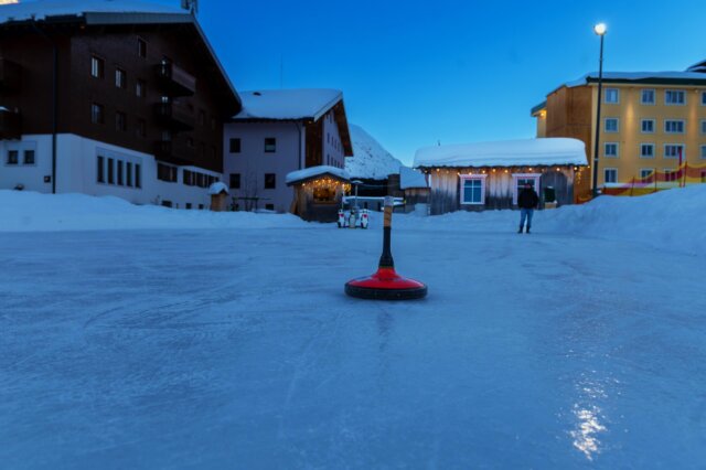 Eisfläche vor dem Hotel Edelweiss in Lech/Zürs mit einem Curlingstein und schneebedeckten Gebäuden im Hintergrund.