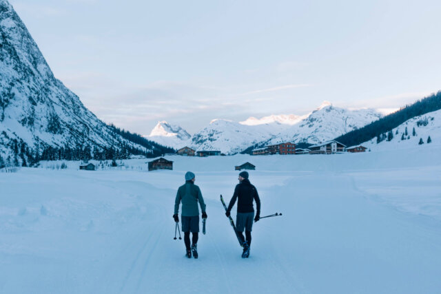 Zwei Personen mit Skiausrüstung gehen durch verschneite Landschaft in Lech/Zürs, umgeben von majestätischen Bergen.