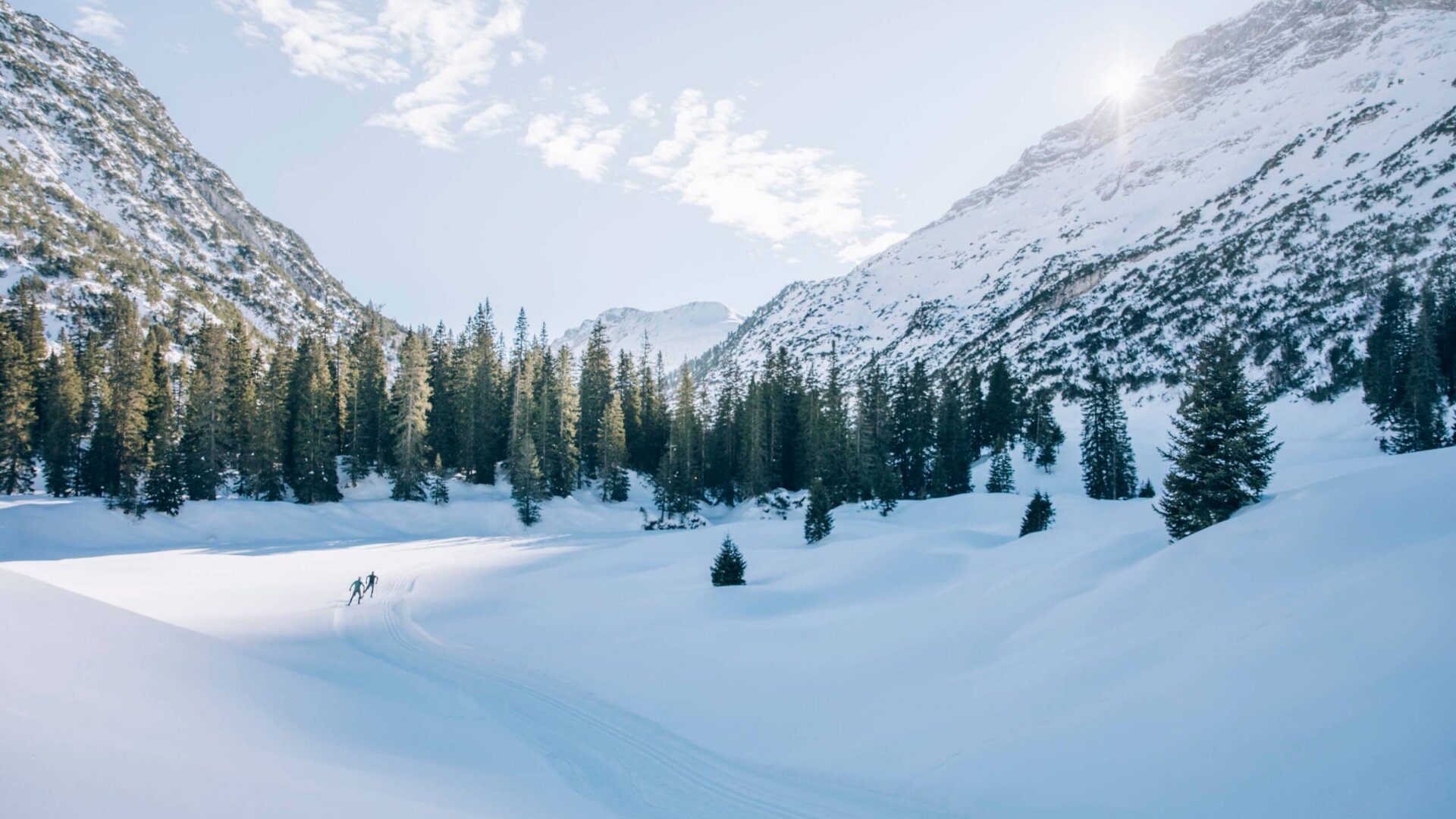 Winterlandschaft in Lech/Zürs mit Skifahrern im Schnee, umgeben von hohen Bergen und verschneiten Tannenbäumen.