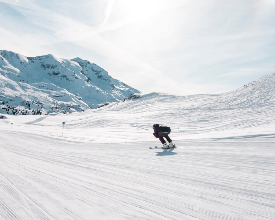 Skifahrer auf einer sonnigen Piste in Lech/Zürs, umgeben von schneebedeckten Bergen und makellosem Himmel.