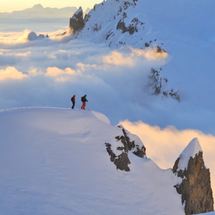 Zwei Skifahrer stehen auf einem schneebedeckten Gipfel über den Wolken im Arlberg, umgeben von atemberaubender Berglandschaft.