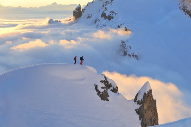 Zwei Skifahrer stehen auf einem schneebedeckten Gipfel über den Wolken im Arlberg, umgeben von atemberaubender Berglandschaft.