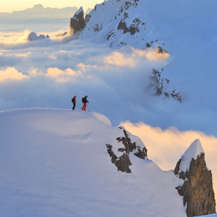 Zwei Skifahrer stehen auf einem schneebedeckten Gipfel über den Wolken im Arlberg, umgeben von atemberaubender Berglandschaft.