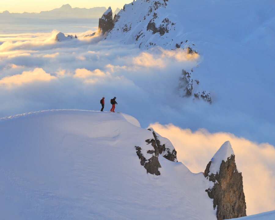 Zwei Skifahrer stehen auf einem schneebedeckten Gipfel über den Wolken im Arlberg, umgeben von atemberaubender Berglandschaft.