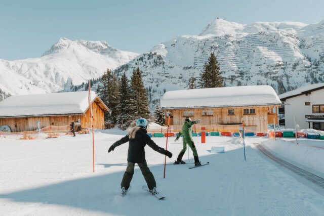 Skifahrer im Winterparadies vor dem Hotel Edelweiss in Lech/Zürs, umgeben von schneebedeckten Bergen und traumhaften Hütten.