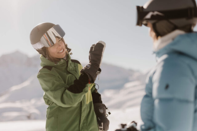 Zwei Frauen im grünen und blauen Skianzug genießen den sonnigen Tag im Schnee in Lech/Zürs. Perfect for winter adventures!