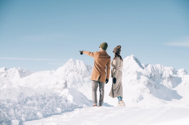 Paar im Winterurlaub betrachtet die Alpenlandschaft in Lech/Zürs am Arlberg, umgeben von schneebedeckten Bergen.