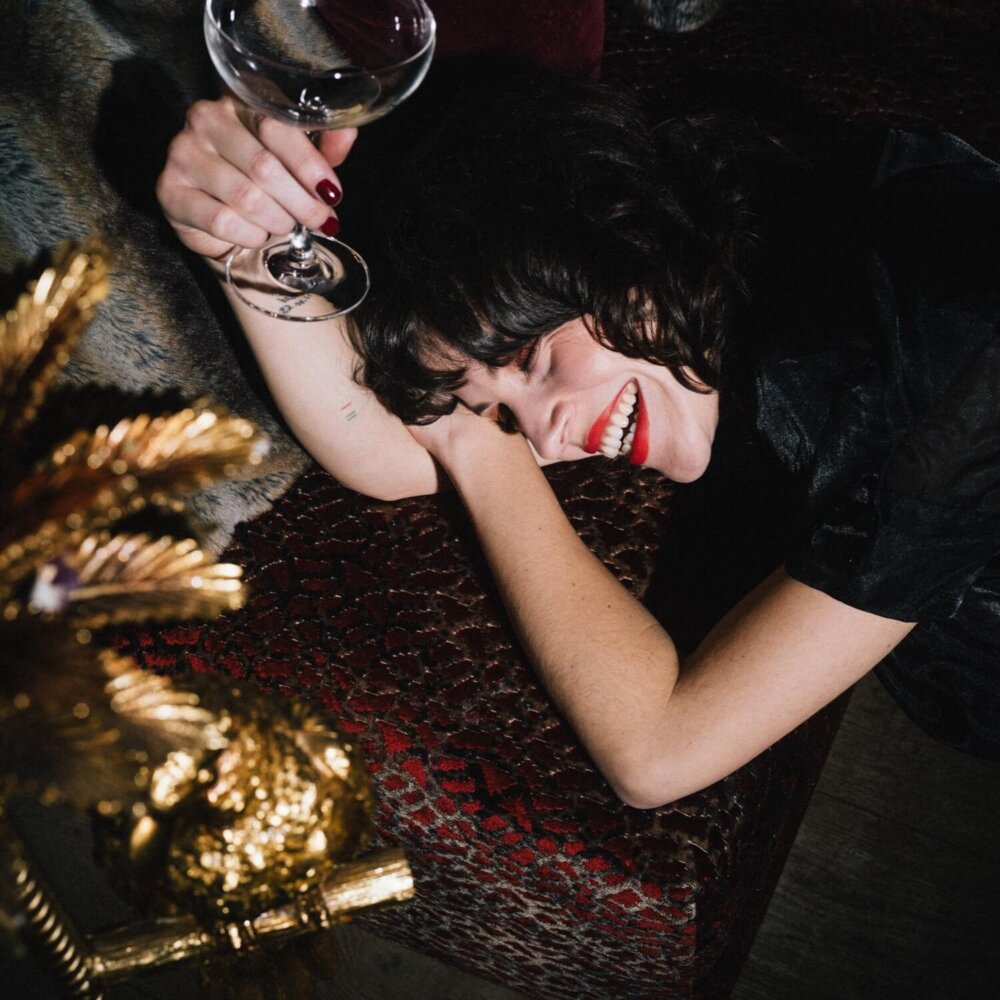 Fröhliche Frau mit Cocktailscheibe auf rotem Sofa im Hotel Edelweiss, Lech/Zürs am Arlberg. Glamouröse Atmosphäre.