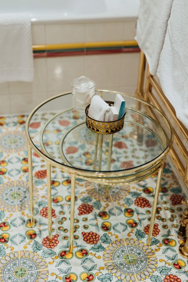 Eleganter Glastisch im Badezimmer des Hotel Edelweiss mit buntem, floralen Fliesenboden und frischen Handtüchern.