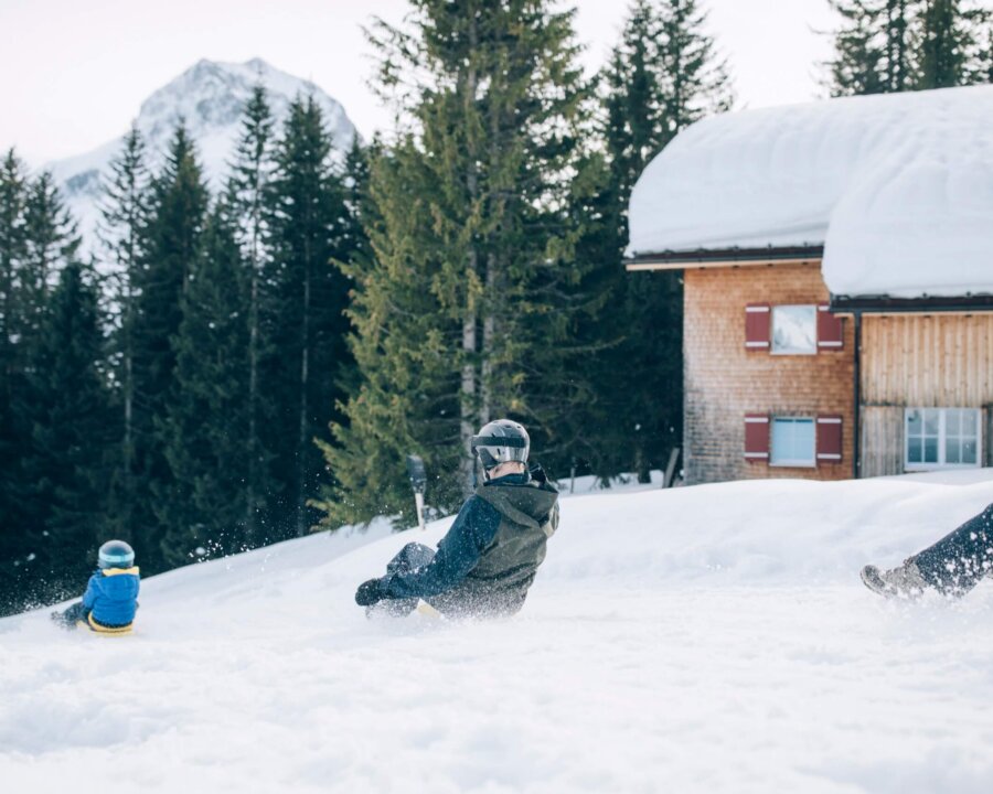 Familie hat Spaß beim Schlittenfahren im Schnee vor dem Hotel Edelweiss in Lech/Zürs, umgeben von winterlicher Natur.