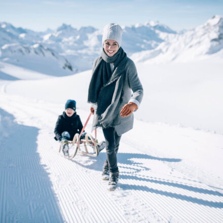 Frau zieht im Schnee einen Schlitten mit einem Kind, umgeben von wunderschöner Winterlandschaft in Lech/Zürs.