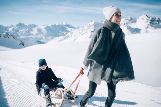 Frau zieht kind auf einem Schlitten durch die schneebedeckte Landschaft in Lech/Zürs am Arlberg. Winterspaß für Familien.