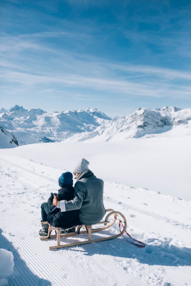 Mutter und Kind auf einem Schlitten im verschneiten Lech, mit atemberaubendem Bergpanorama im Hintergrund.