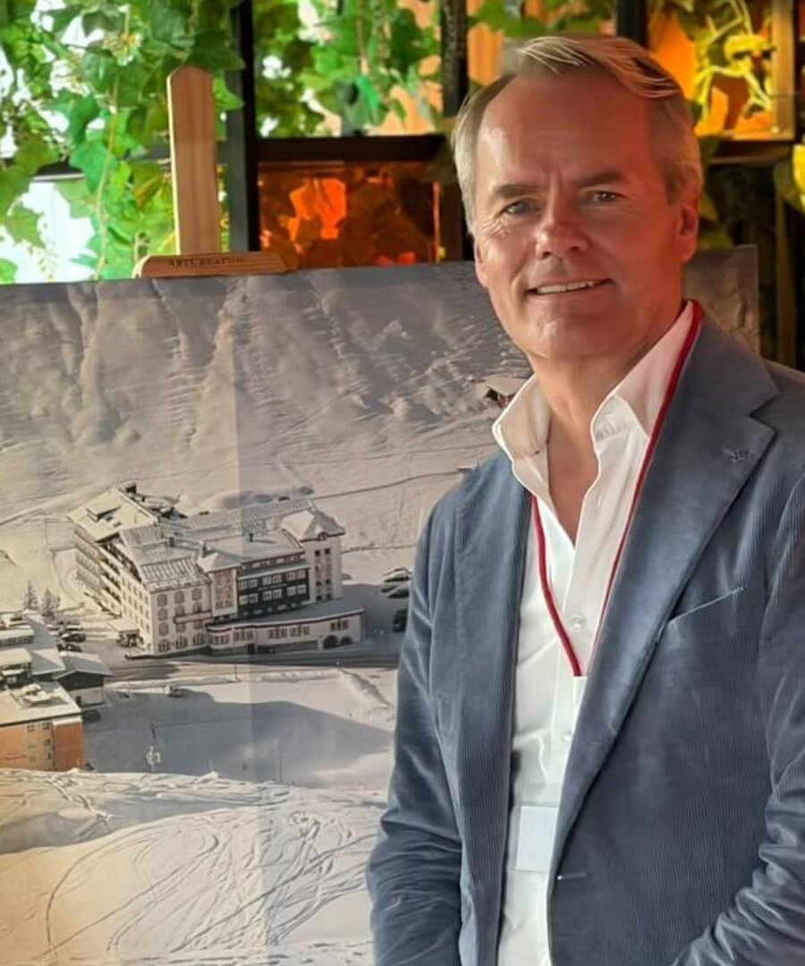 Mann mit lächelndem Gesicht steht vor einem winterlichen Bild des Hotel Edelweiss in Lech/Zürs am Arlberg.